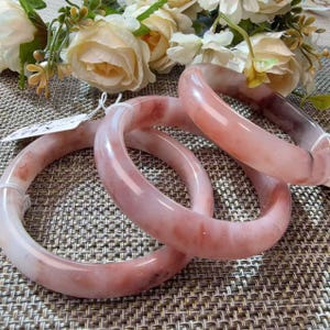 Puede incluir: Tres brazaletes rígidos lisos y pulidos, hechos de una piedra translúcida de color rosa pálido con vetas de color marrón rojizo más oscuro. Cada brazalete circular presenta un perfil redondeado y un acabado brillante, mostrando variaciones de color naturales.