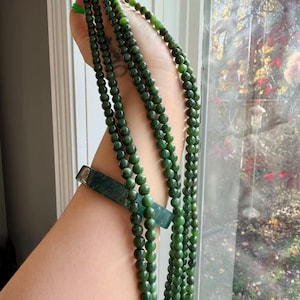 May include: A long strand of dark green, spherical jade beads featuring a smooth, polished finish. The necklace is draped in multiple loops, showcasing a consistent deep forest green color throughout the circular stone beads.