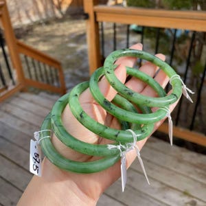 May include: Four jade bangles in varying shades of green are held in a hand. The bangles have a smooth, polished finish with dark flecks. Each bangle has a tag attached with the number 57.5.