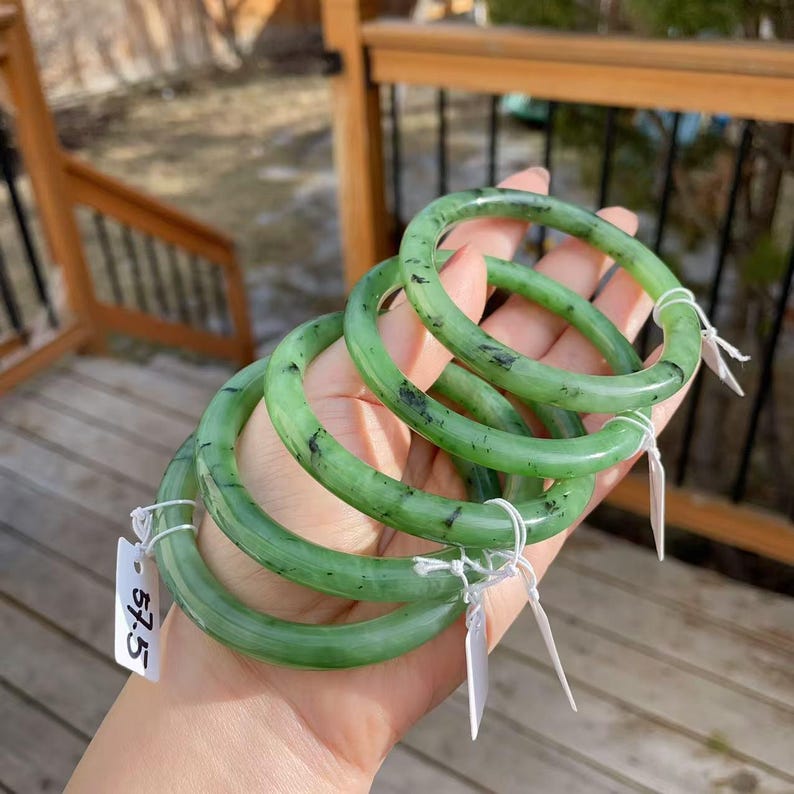 May include: Four jade bangles in varying shades of green are held in a hand. The bangles have a smooth, polished finish with dark flecks. Each bangle has a tag attached with the number 57.5.