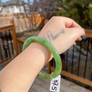 May include: A light green jade bangle bracelet. The bracelet is a solid, smooth band of translucent green stone. The bracelet is worn on a wrist. A white tag with the number 57.5 is attached to the bracelet.