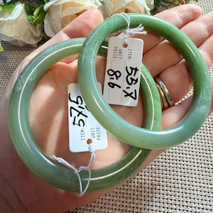 Peut inclure: Deux bracelets joncs en jade vert, lisses et polis. Chaque bracelet est muni d'une petite étiquette rectangulaire blanche attachée par un cordon blanc. Une étiquette porte le nombre 53.7 écrit à la main, l'autre porte le nombre 57.5.
