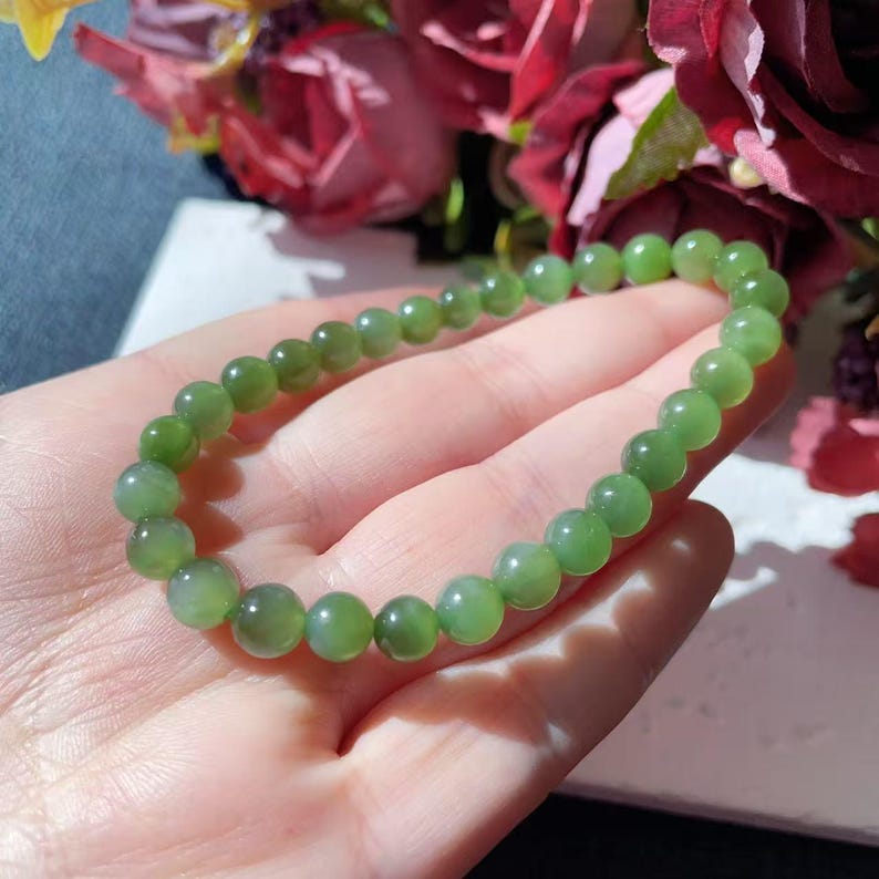 May include: A green jade bead bracelet. The bracelet features round, polished beads in various shades of green. The beads are strung together to create a flexible band. The bracelet is displayed against a backdrop of flowers.