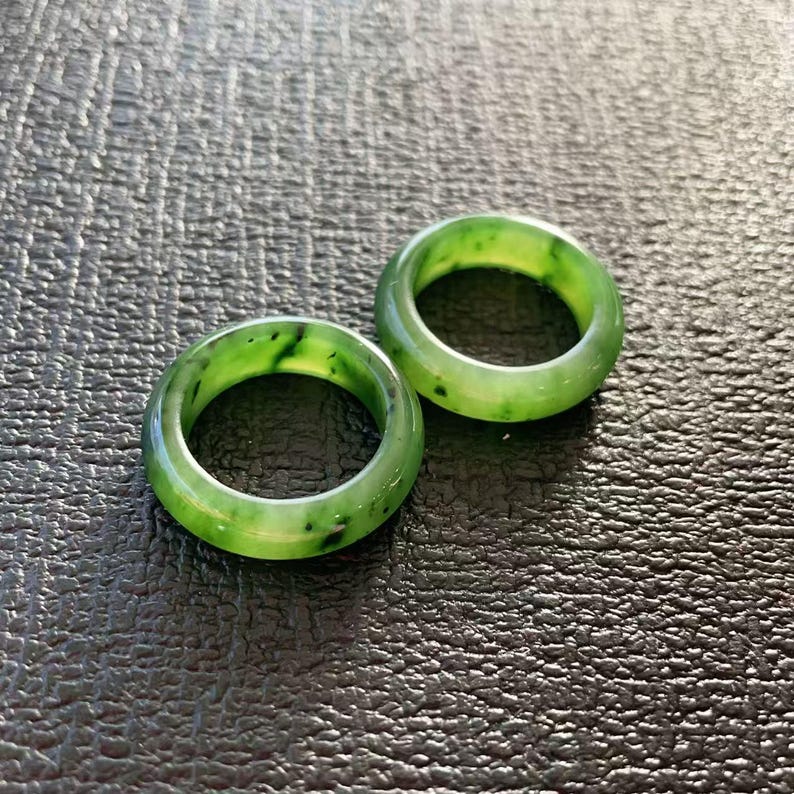 May include: Two jade rings with a vibrant green hue and dark green speckles. The rings are circular and smooth, set against a dark, textured surface. The rings are a classic jewelry item.