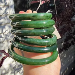 May include: A stack of five smooth, polished green jade bangles with varying shades of deep forest green and translucent emerald tones. Each circular bracelet features a rounded profile and a glossy, reflective surface, showcasing natural color variations.