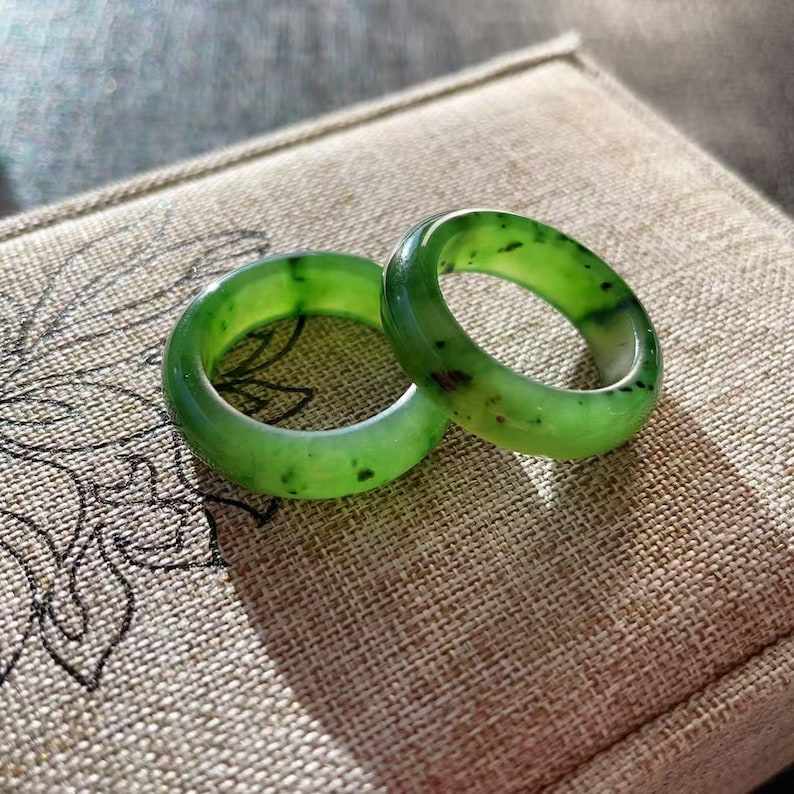 May include: Two jade rings with a vibrant green hue and dark speckles. The rings are smooth and polished, resting on a textured, tan surface. The rings are a classic band style, perfect for everyday wear.