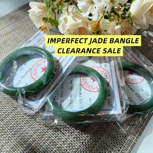 May include: Three dark green jade bangles, each individually wrapped in clear plastic packaging and accompanied by a white certificate of authenticity with red circular stamps and black text. The text IMPERFECT JADE BANGLE CLEARANCE SALE is visible.