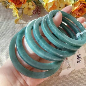 May include: A stack of five smooth, translucent, sage green jade-colored bangles. The bracelets are circular with a rounded profile. A white paper tag is attached to the bangles with white string, featuring the handwritten text 56.5.