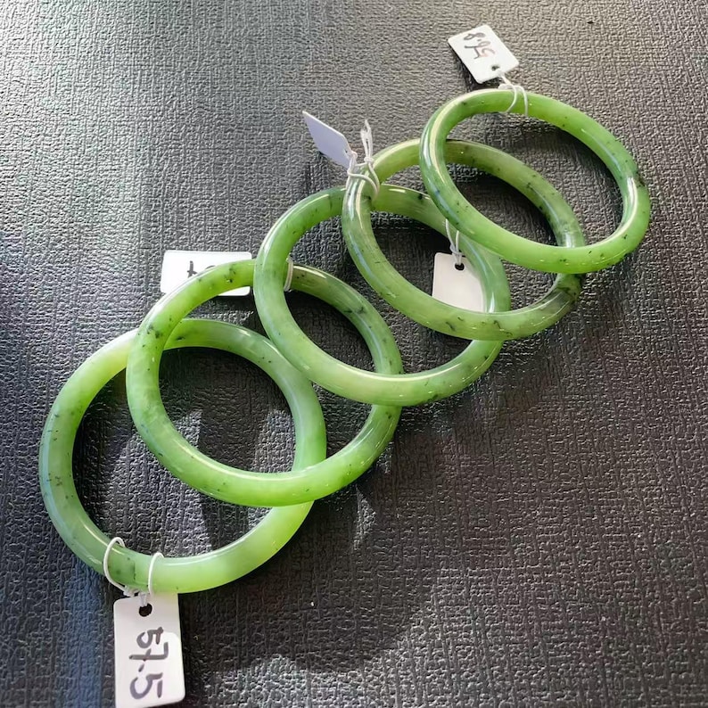 May include: Five jade bangles in varying shades of green are displayed on a dark surface. Each bangle is a smooth, circular band with a polished finish. Price tags are attached to some of the bangles.