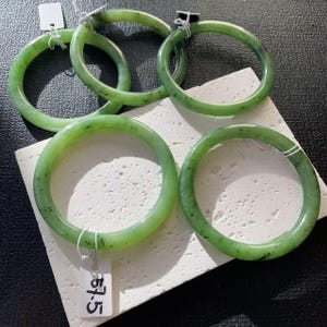 May include: A collection of five jade bangles in varying sizes. The bangles are a translucent green color with darker green speckles. Each bangle is round and smooth, with a polished finish. One bangle has a tag that reads "57.5".