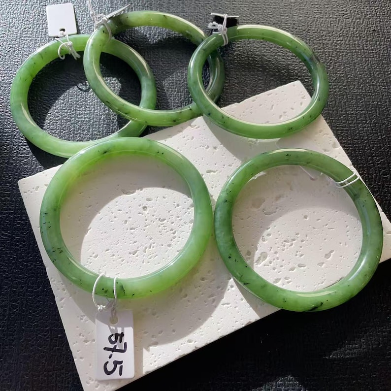 May include: A collection of five jade bangles in varying sizes. The bangles are a translucent green color with darker green speckles. Each bangle is round and smooth, with a polished finish. One bangle has a tag that reads "57.5".