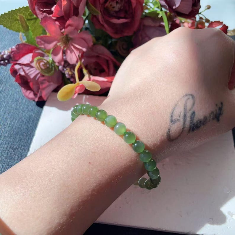 May include: A green beaded bracelet. The bracelet features round, polished beads in shades of green. The bracelet is worn on a wrist, with a tattoo of the word "Phoenix" visible.