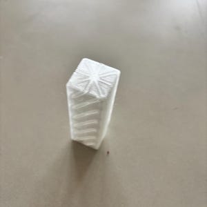 Könnte beinhalten: Ein weißes, geometrisches 3D-gedrucktes Objekt mit einem Sternmuster auf der Oberseite und horizontalen Linien an den Seiten. Das Objekt ist rechteckig mit leicht abgewinkelter Ober- und Unterseite.