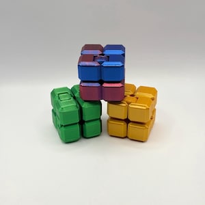 May include: Three colourful fidget cubes in green, gold, and a combination of blue and red. Each cube is made of multiple connected blocks, designed for tactile play and stress relief. The cubes are arranged on a white surface.