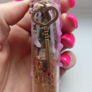 May include: A small glass vial filled with dried herbs, crystals, and a bronze-coloured key charm. The vial is sealed with a twine-wrapped cork and pink wax. The key charm has a heart-shaped top.