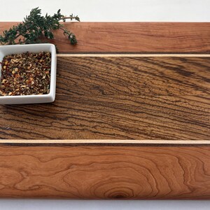 May include: A rectangular wooden cutting board featuring a wide central panel of dark brown wood with prominent grain, flanked by two lighter reddish-brown wood sections and thin white inlay strips. A small white square bowl holds dried spices.