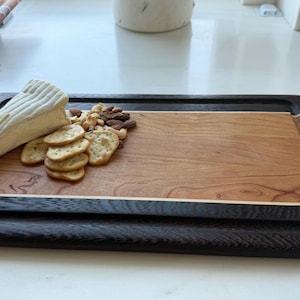 May include: A rectangular wooden serving board featuring a light brown center panel framed by dark brown wood borders. A wedge of white brie cheese, a small pile of round crackers, and mixed nuts are arranged on the left side with a gold cheese knife.