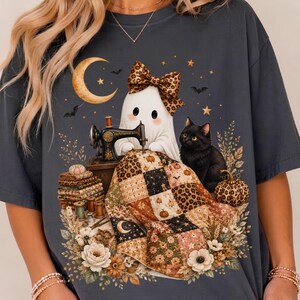 Può includere: Una t-shirt grigio scuro con la grafica di un fantasma bianco che indossa un fiocco leopardato, seduto a una macchina da cucire vintage. Un gatto nero siede accanto a una trapunta patchwork, zucche, fiori, una luna crescente e piccoli pipistrelli.