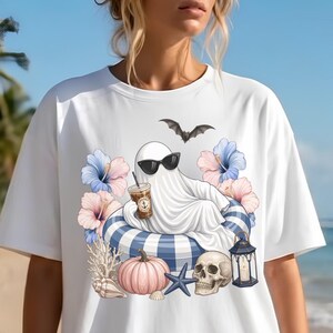 Op de afbeelding: Wit T-shirt met een cartoon spook met een zonnebril, ontspannen in een blauw-witte gestreepte zwemband, met een koffie. Het ontwerp bevat een vleermuis, bloemen, een pompoen, een schedel en een lantaarn.