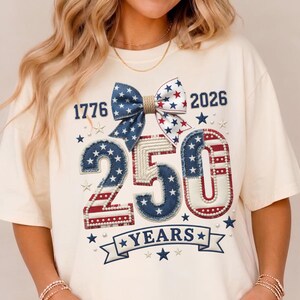 Op de afbeelding: Crèmekleurig T-shirt met een patriottisch ontwerp. Voorzien van de cijfers "1776" en "2026" die een strik met sterpatronen flankeren. Het nummer "250" is prominent weergegeven, samen met het woord "YEARS" op een banner, allemaal in een patriottisch thema.