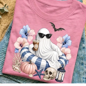 Op de afbeelding: Roze t-shirt met een spook met een zonnebril, drijvend op een blauw-wit gestreepte zwemband, met een koffie in de hand. Het ontwerp bevat een vleermuis, bloemen, een pompoen, een schedel en een lantaarn.