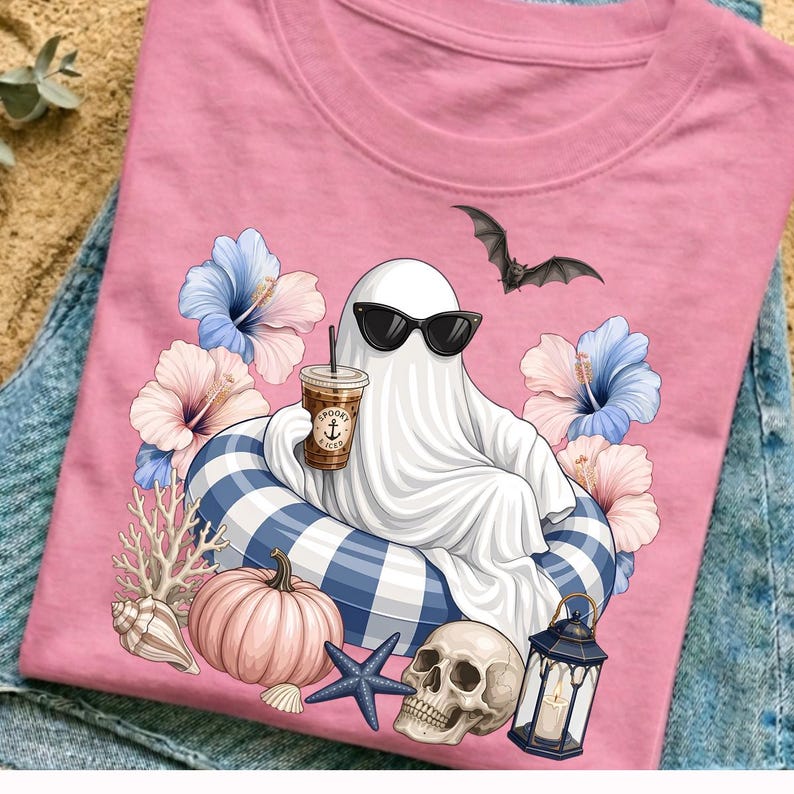 Op de afbeelding: Roze t-shirt met een spook met een zonnebril, drijvend op een blauw-wit gestreepte zwemband, met een koffie in de hand. Het ontwerp bevat een vleermuis, bloemen, een pompoen, een schedel en een lantaarn.