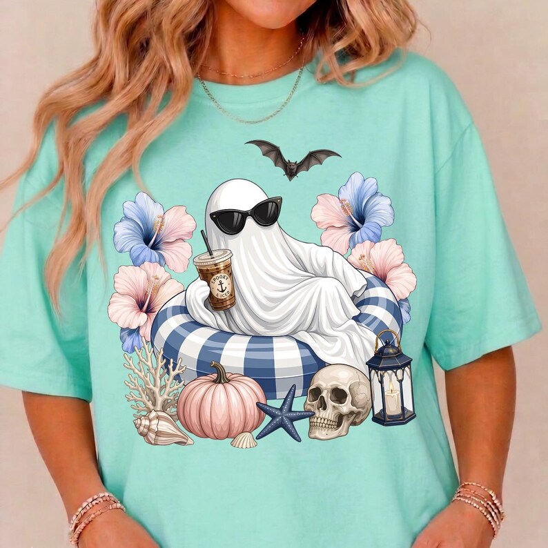 Op de afbeelding: Lichtblauwgroen T-shirt met een cartoongeest met een zonnebril, ontspannen op een blauw-wit gestreepte vlot, met een kopje koffie. Het ontwerp bevat bloemen, een vleermuis, een pompoen, een schedel en schelpen.