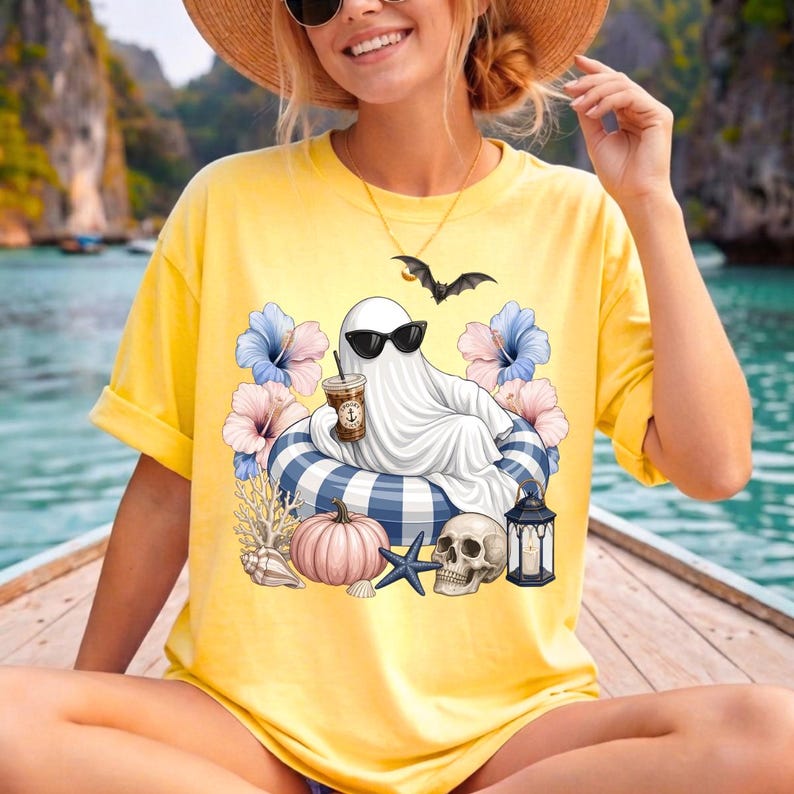 Op de afbeelding: Geel t-shirt met een cartoongeest met een zonnebril, ontspannen op een blauw-wit gestreepte zwemband, met een koffie. Het ontwerp bevat bloemen, een pompoen, een schedel en een vleermuis.