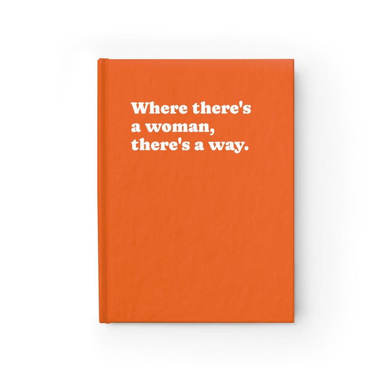 Op de afbeelding: Een feloranje notitieboek met harde kaft en de witte tekst "Where there's a woman, there's a way." op de voorkant. Het notitieboek heeft een effen, levendige oranje achtergrond en een strak, rechthoekig ontwerp met een verticale rug.