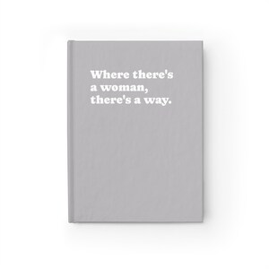 Op de afbeelding: Een lichtgrijs notitieboek met harde kaft en de in het wit gedrukte tekst "Where there's a woman, there's a way." op de voorkant. Het notitieboek heeft een strak, effen oppervlak met een verticale rug aan de linkerkant van het object.