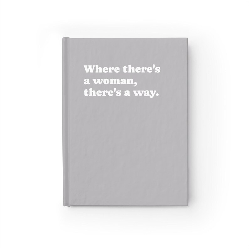 Op de afbeelding: Een lichtgrijs notitieboek met harde kaft en de in het wit gedrukte tekst "Where there's a woman, there's a way." op de voorkant. Het notitieboek heeft een strak, effen oppervlak met een verticale rug aan de linkerkant van het object.