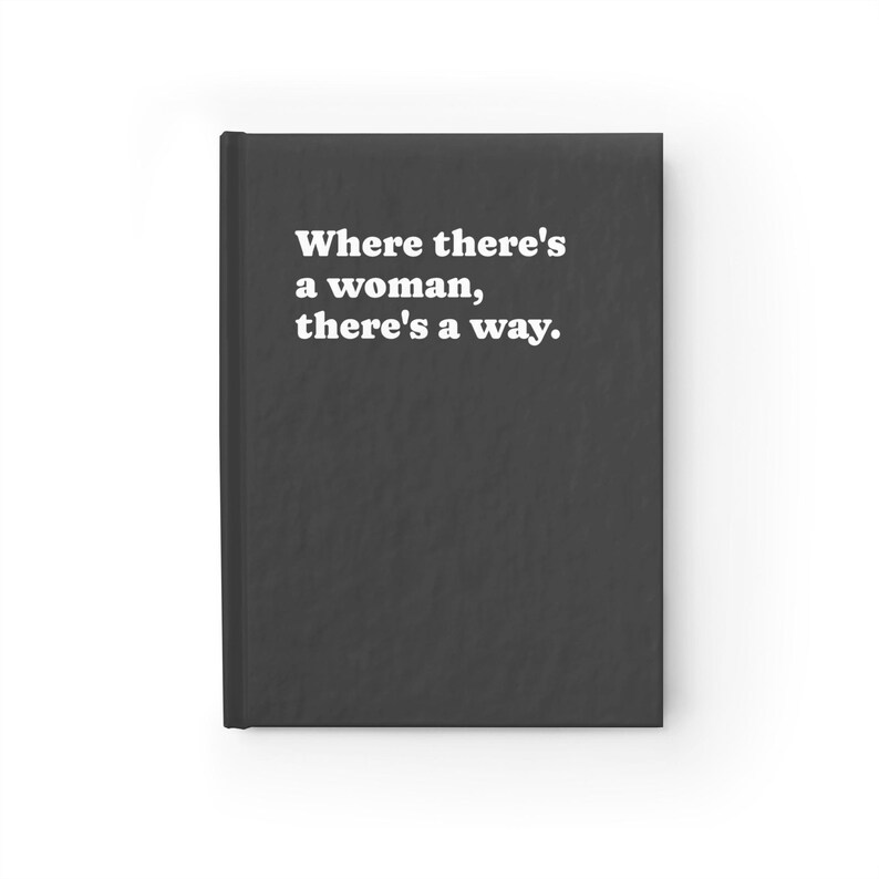 Op de afbeelding: Een zwart notitieboek met harde kaft en de in het wit gedrukte tekst Where there's a woman, there's a way. Het notitieboek heeft een effen donkergrijs-zwarte kaft met een matte afwerking en een verticale rug aan de linkerkant van de voorkant.