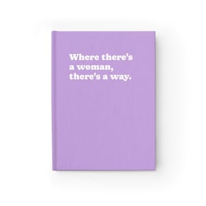Op de afbeelding: Een lichtpaars notitieboek met harde kaft en de witte tekst "Where there's a woman, there's a way." op de voorkant. Het notitieboek heeft een effen lavendelkleurige achtergrond en een verticale rug aan de linkerkant van de kaft.