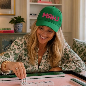 May include: A green baseball cap with the word MAHJ embroidered in bright pink letters on the front. The cap features a structured crown, a curved brim, and an adjustable strap at the back, providing a classic fit for various head sizes.