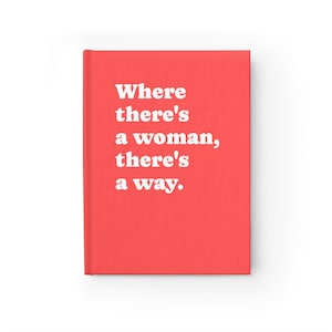 Op de afbeelding: Een fel koraalkleurig notitieboek met harde kaft en de witte tekst "Where there's a woman, there's a way." op de voorkant. Het notitieboek heeft een strakke, effen achtergrond en een verticale rug aan de linkerkant van de omslag.