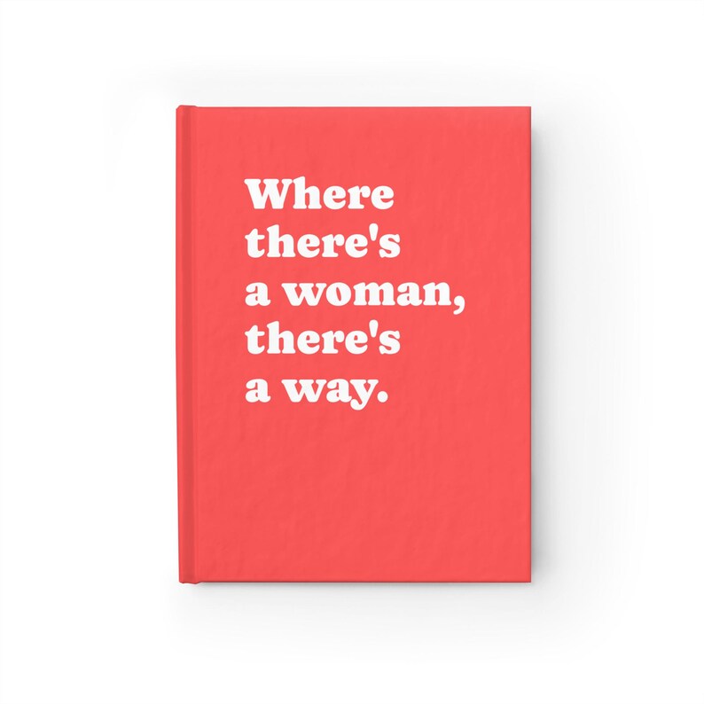 Op de afbeelding: Een fel koraalkleurig notitieboek met harde kaft en de witte tekst "Where there's a woman, there's a way." op de voorkant. Het notitieboek heeft een strakke, effen achtergrond en een verticale rug aan de linkerkant van de omslag.