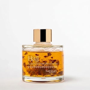 Peut inclure: Un flacon en verre transparent rempli d'un liquide ambré et de morceaux de plantes. L'étiquette porte le texte Hajj, A sacred journey like no other, et barakah home. Le flacon est surmonté d'un bouchon cylindrique en métal doré.