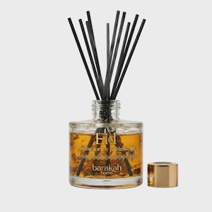 Può includere: Un flacone diffusore in vetro trasparente con bastoncini neri e un tappo dorato. La bottiglia contiene un liquido dorato con elementi decorativi e il testo "Eid A time for joy & blessings" e "barakah home".