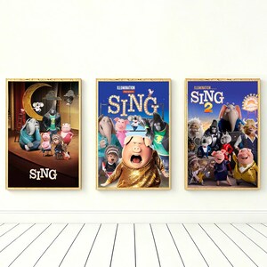 Könnte beinhalten: Drei gerahmte Filmplakate der Animationsfilme "Sing" und "Sing 2". Die Poster zeigen farbenfrohe Illustrationen verschiedener Tierfiguren. Die Poster sind in goldenen Rahmen und werden vor einer weißen Wand ausgestellt.