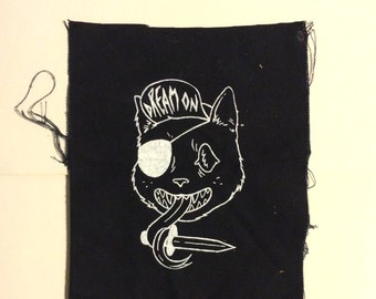 May include: Black and white embroidered patch of a cat wearing a hat that says "Dream On" and an eyepatch, holding a dagger in its mouth.