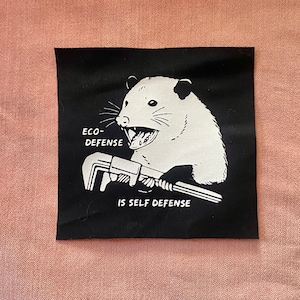 May include: Black and white patch featuring a cartoon possum holding a wrench. The text on the patch reads "Eco-Defense is Self Defense."