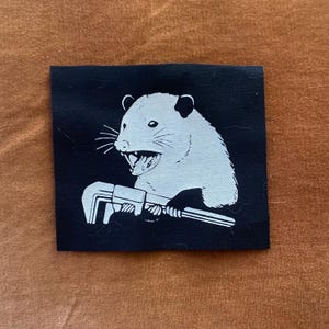 May include: Black and white iron-on patch featuring a cartoon opossum with an open mouth and a wrench in its hand.