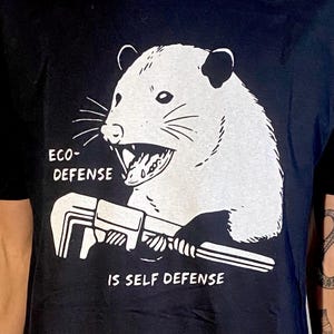 May include: Black t-shirt with a white illustration of an opossum holding a wrench. The text "ECO-DEFENSE IS SELF DEFENSE" is printed below the image.