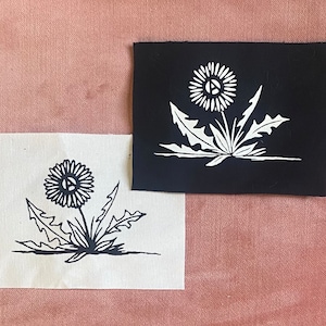 Könnte beinhalten: Zwei schwarze und weiße Stoffpatches mit einem Löwenzahnmuster. Der Löwenzahn ist auf einem Patch weiß auf schwarzem Hintergrund und auf dem anderen Patch schwarz auf weißem Hintergrund umrandet.