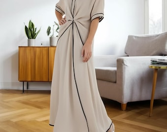 May include: A beige maxi dress with black trim and a V-neckline. The dress has short sleeves and a knot detail at the waist. The dress is worn with white sandals. The dress is suitable for casual or semi-formal occasions.