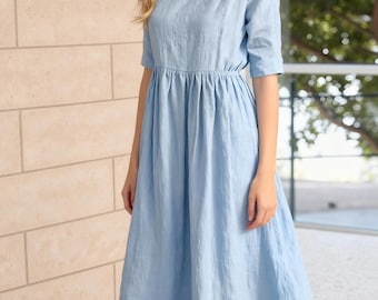 May include: A pale blue dress with short sleeves and a gathered waist. The dress has a round neckline and a long, flowing skirt. The fabric looks like a lightweight linen or cotton blend, suitable for warmer weather.