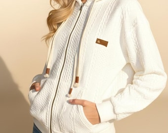 May include: A cream-coloured zip-up hoodie with a textured knit pattern. The hoodie has a hood with drawstrings, front pockets, and a small brown leather patch. The model is wearing blue jeans.