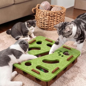 以下が含まれることがあります： 3匹の猫が、複数の開口部と迷路のようなデザインの緑と茶色の猫用おもちゃで遊んでいます。背景には毛糸玉が入ったバスケットがあります。猫たちはベージュ色のカーペットの上でおもちゃと遊んでいます。