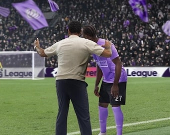 Puede incluir: Un gráfico digital con el texto FOOTBALL MANAGER 27. Debajo, un entrenador con camisa beige y pantalón oscuro está en un campo de fútbol verde, poniendo una mano sobre el hombro de un jugador con camiseta morada, pantalón corto negro y medias moradas.