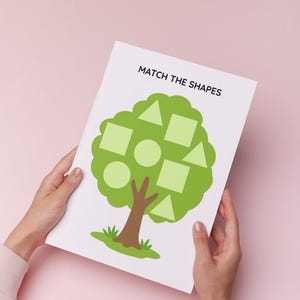 May include: A white rectangular activity sheet featuring the text MATCH THE SHAPES at the top. The centre displays a green tree illustration with various light green geometric shapes including triangles, squares, and circles integrated into the foliage.